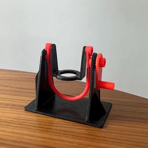 May include: A black plastic 3D printed device with a red rubber band and red plastic pieces. The device is designed to hold a small object in place.