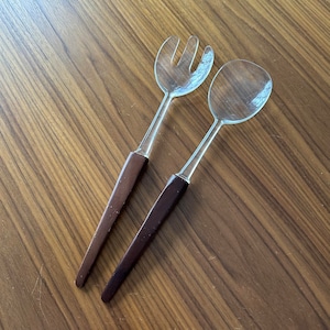 May include: A set of two clear plastic serving utensils with brown wooden handles. The fork has four tines and the spoon has a rounded bowl.