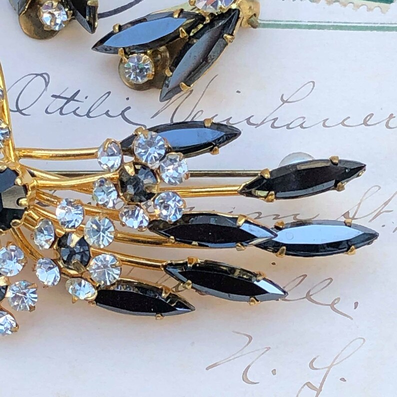 Puede incluir: Un broche de tono dorado con diamantes de imitaci&oacute;n negros y transparentes. El broche tiene forma de abanico con m&uacute;ltiples puntas. Los diamantes de imitaci&oacute;n est&aacute;n engastados en un metal de tono dorado.