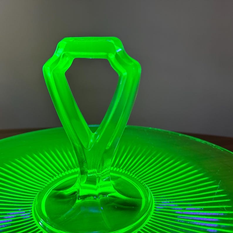 Może przedstawiać: Zbliżenie jasnoniebieskiego szklanego p&oacute;łmiska do serwowania z uchwytem. Uchwyt ma geometryczny kształt. Taca ma okrągłą podstawę z wzorem promieni słonecznych promieniujących ze środka.