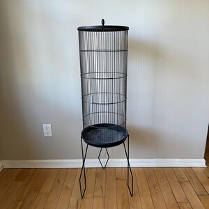 May include: Black metal bird cage with a round base and a stand with four legs. The cage is tall and cylindrical.