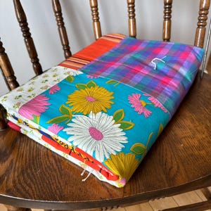 May include: A patchwork quilt with a variety of colorful fabrics, including floral patterns, plaid, and stripes. The quilt is folded and placed on a wooden chair.