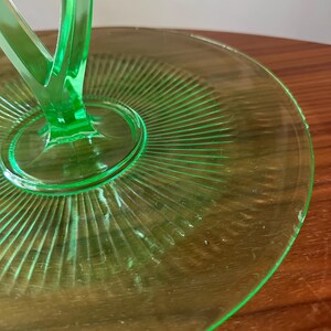 Może przedstawiać: Zbliżenie na zabytkowy zielony szklany p&oacute;łmisek. Naczynie ma okrągły talerz z żebrowanym wzorem promieniującym ze środka. Centralny trzon podtrzymuje uchwyt. Szkło ma jasny, p&oacute;łprzezroczysty wygląd.