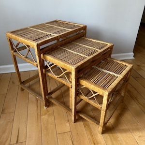 May include: A set of three nested wicker tables with a natural brown finish. The tables are made of woven rattan and have a square shape. The tables are stacked on top of each other, with the largest table at the bottom and the smallest table at the top.