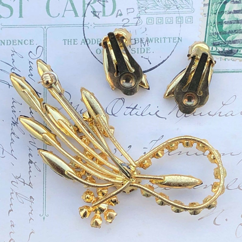 Puede incluir: Un broche de tono dorado con un racimo de diamantes de imitaci&oacute;n en forma de flor. El broche est&aacute; unido a un juego de pendientes a juego con cierre de clip.