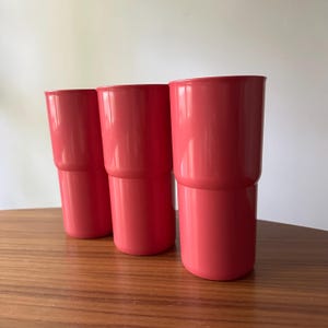 May include: Three coral-colored plastic tumblers are stacked on a wooden surface. The tumblers have a two-part design, with a slightly smaller base section and a wider top. The background is a plain white wall.