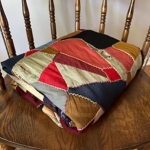 May include: A folded patchwork quilt made from various fabrics in shades of red, blue, brown, and green. The quilt is made of squares and triangles sewn together with a contrasting thread.