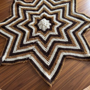 May include: A brown, white, and tan crocheted star-shaped blanket. The blanket is made with a repeating pattern of stripes.