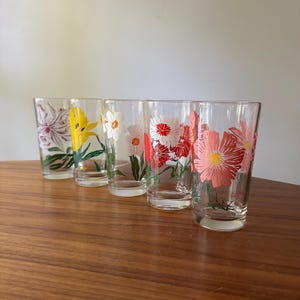 May include: Five clear drinking glasses with floral designs. Each glass features a different flower in shades of pink, red, yellow, and white, with green stems and leaves. The glasses are arranged on a wooden surface.