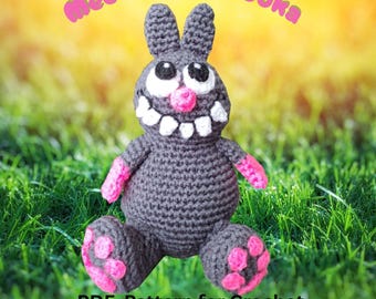 May include: A crocheted gray amigurumi bunny with pink accents, including paws and arms. The bunny has a wide, toothy grin and large eyes. The text "Meet Muff Pooka" and "PDF Pattern for Crochet 10" Inch Amigurumi" are visible.