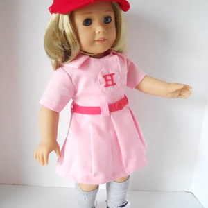 May include: A doll wearing a pink baseball uniform with a red cap and a red "H" on the front. The doll is wearing grey socks and black and white sneakers.