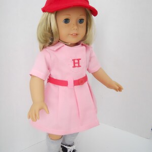 May include: A doll wearing a pink dress with a red belt and a red baseball cap with a white "H" on it. The doll has blonde hair and blue eyes. The doll is wearing grey socks and black and white sneakers.
