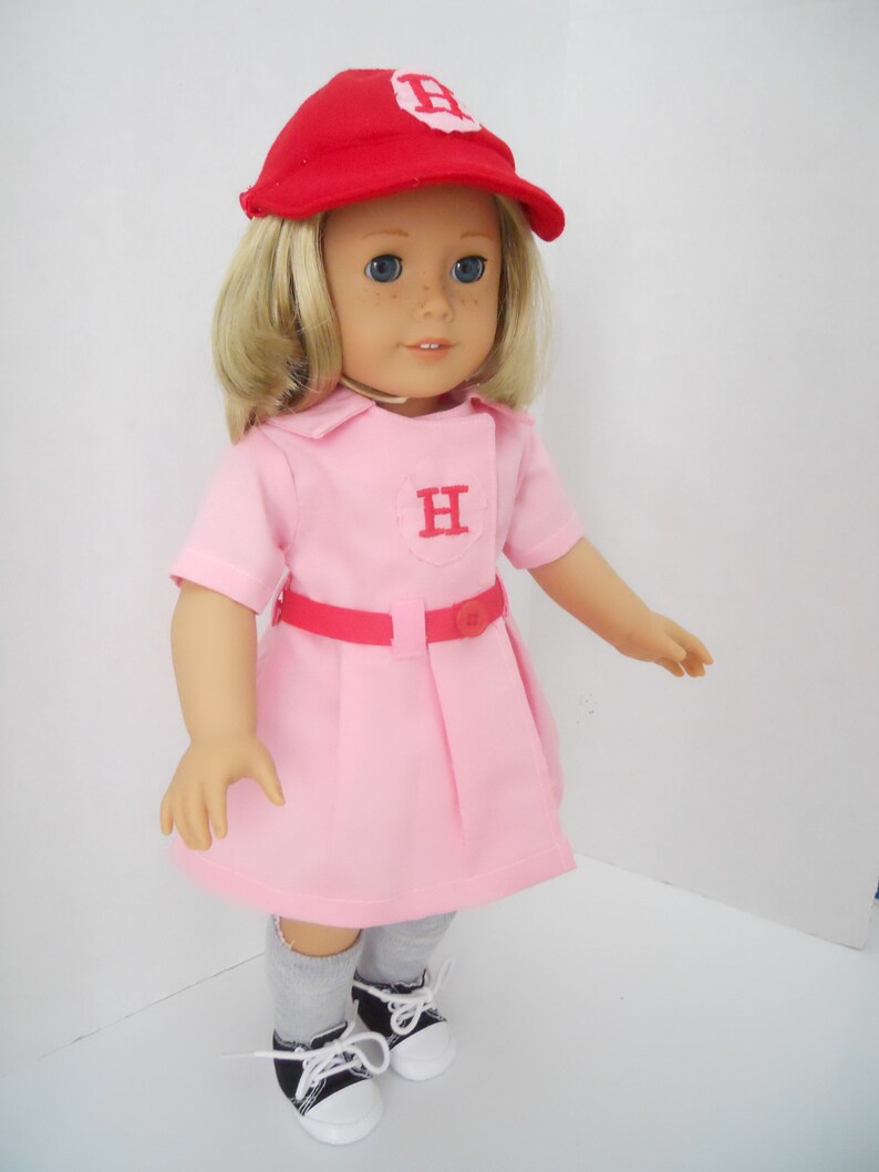 May include: A doll wearing a pink dress with a red belt and a red baseball cap with a white "H" on it. The doll has blonde hair and blue eyes. The doll is wearing grey socks and black and white sneakers.