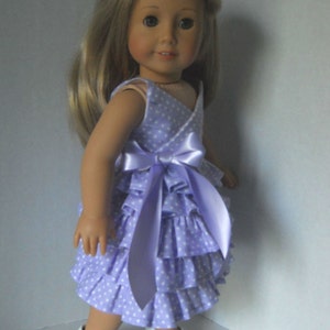 May include: A doll wearing a lavender dress with white polka dots and a matching lavender satin bow. The dress has multiple ruffles and the doll is wearing white shoes.
