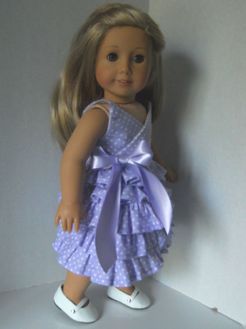 May include: A doll wearing a lavender dress with white polka dots and a matching lavender satin bow. The dress has multiple ruffles and the doll is wearing white shoes.
