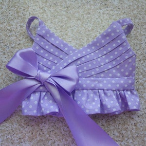 May include: A lavender dress with white polka dots and a matching lavender satin bow. The dress has a ruffled hem and adjustable straps.