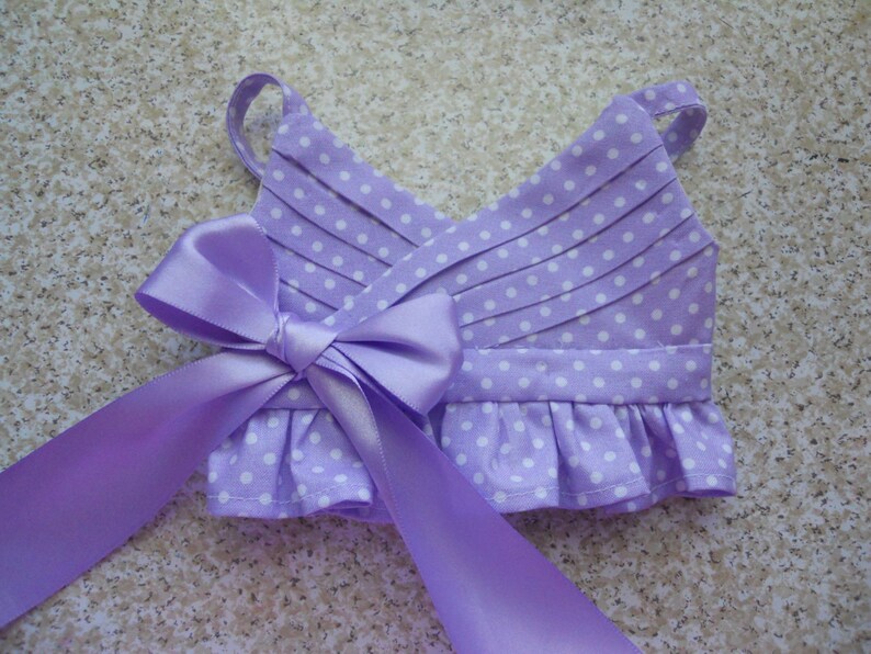 May include: A lavender dress with white polka dots and a matching lavender satin bow. The dress has a ruffled hem and adjustable straps.