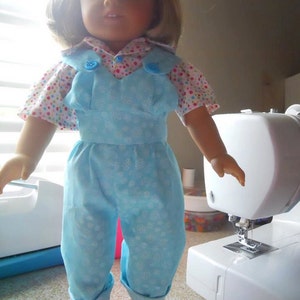 Puede incluir: Una mu&ntilde;eca con un mono azul con estampado de copo de nieve blanco y una camisa de lunares rosa y blanco con botones azules. La mu&ntilde;eca tiene el pelo rubio y los ojos azules.