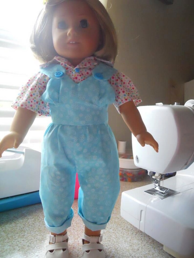 Puede incluir: Una mu&ntilde;eca con un mono azul con estampado de copo de nieve blanco y una camisa de lunares rosa y blanco con botones azules. La mu&ntilde;eca tiene el pelo rubio y los ojos azules.
