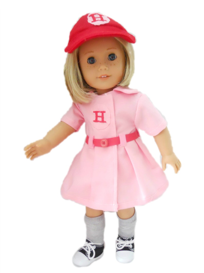 May include: A pink and red baseball uniform for a doll, featuring a red cap with a white "H" on the front, a pink dress with a white "H" on the chest, a red belt, and gray socks with black sneakers.