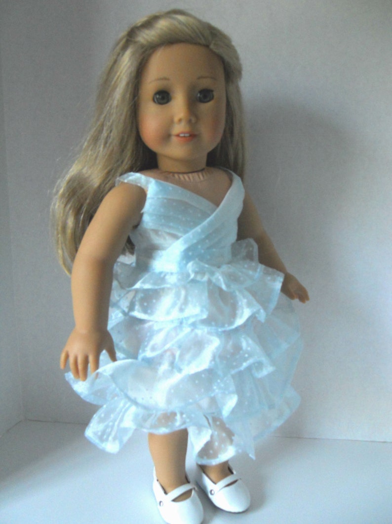 May include: A doll wearing a light blue dress with a ruffled skirt and white shoes.