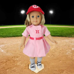 May include: A doll wearing a pink baseball uniform with a red cap and the letter "H" on the front. The doll is standing on a baseball base.