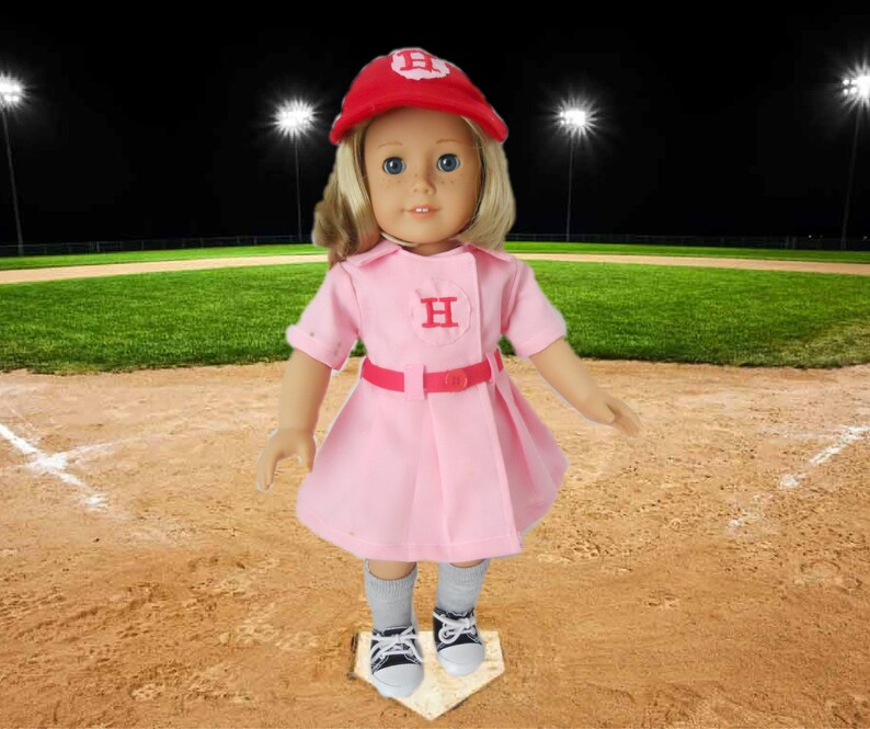 May include: A doll wearing a pink baseball uniform with a red cap and the letter "H" on the front. The doll is standing on a baseball base.