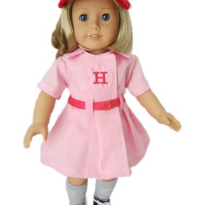May include: A pink and red baseball uniform for a doll, featuring a red cap with a white "H" on it, a pink dress with a white "H" on the chest, and a red belt. The doll is wearing gray socks and black and white sneakers.