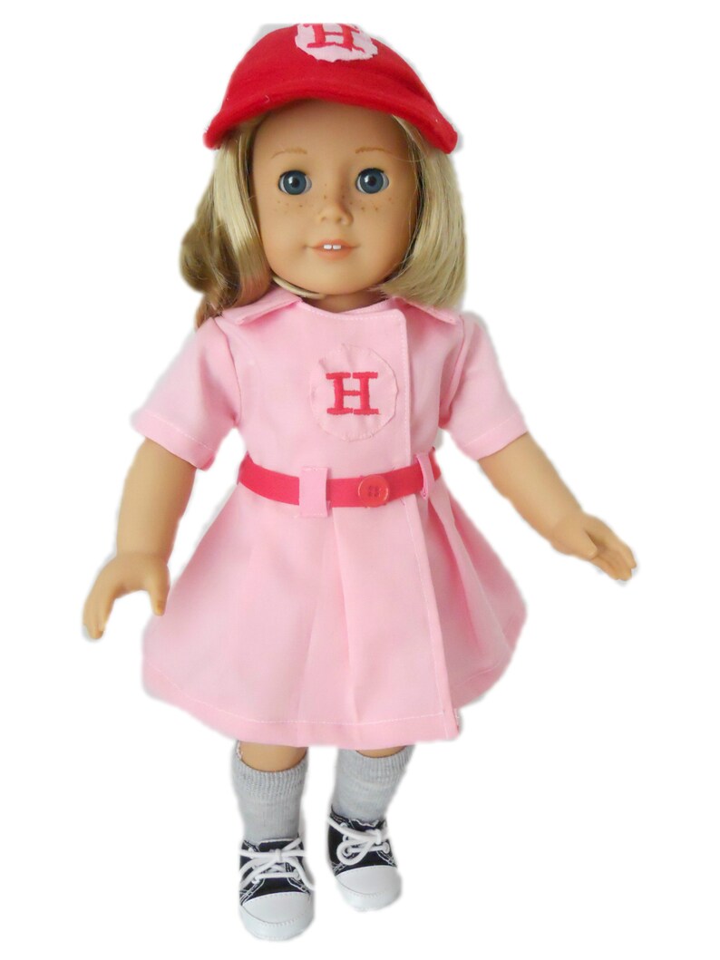 May include: A pink and red baseball uniform for a doll, featuring a red cap with a white "H" on it, a pink dress with a white "H" on the chest, and a red belt. The doll is wearing gray socks and black and white sneakers.