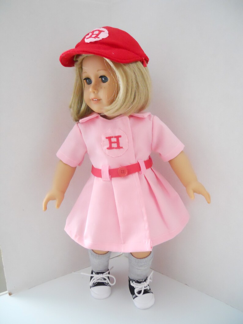May include: A pink dress with a red belt and a red baseball cap with a white "H" on it. The doll is wearing grey socks and black and white sneakers.