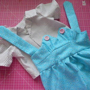 Puede incluir: Un mono azul y blanco con estampado de flores con dos tirantes y botones rosas. El mono est&aacute; sobre una alfombra de corte rosa. Una camisa blanca con lunares negros est&aacute; sobre el mono.