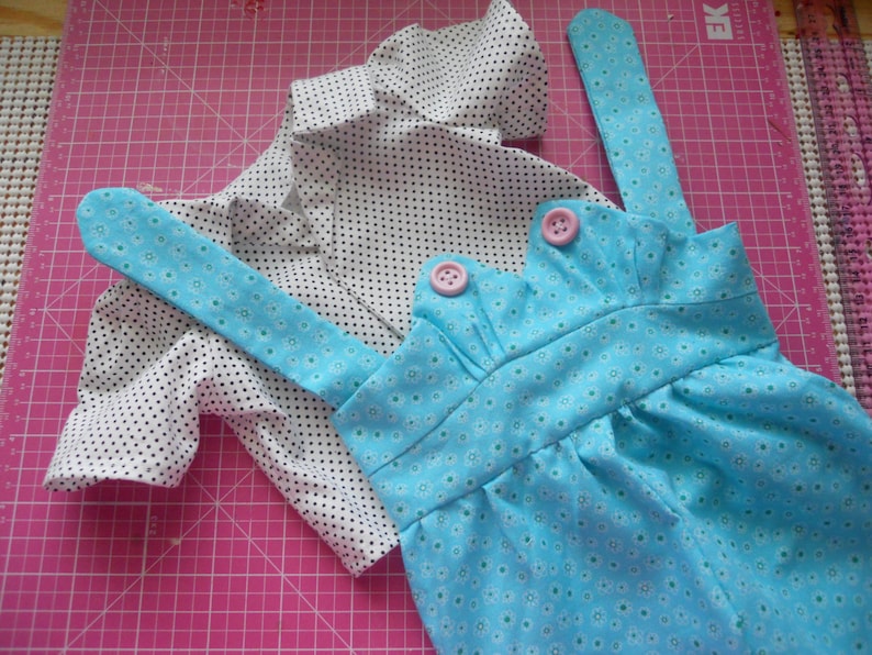 Puede incluir: Un mono azul y blanco con estampado de flores con dos tirantes y botones rosas. El mono est&aacute; sobre una alfombra de corte rosa. Una camisa blanca con lunares negros est&aacute; sobre el mono.