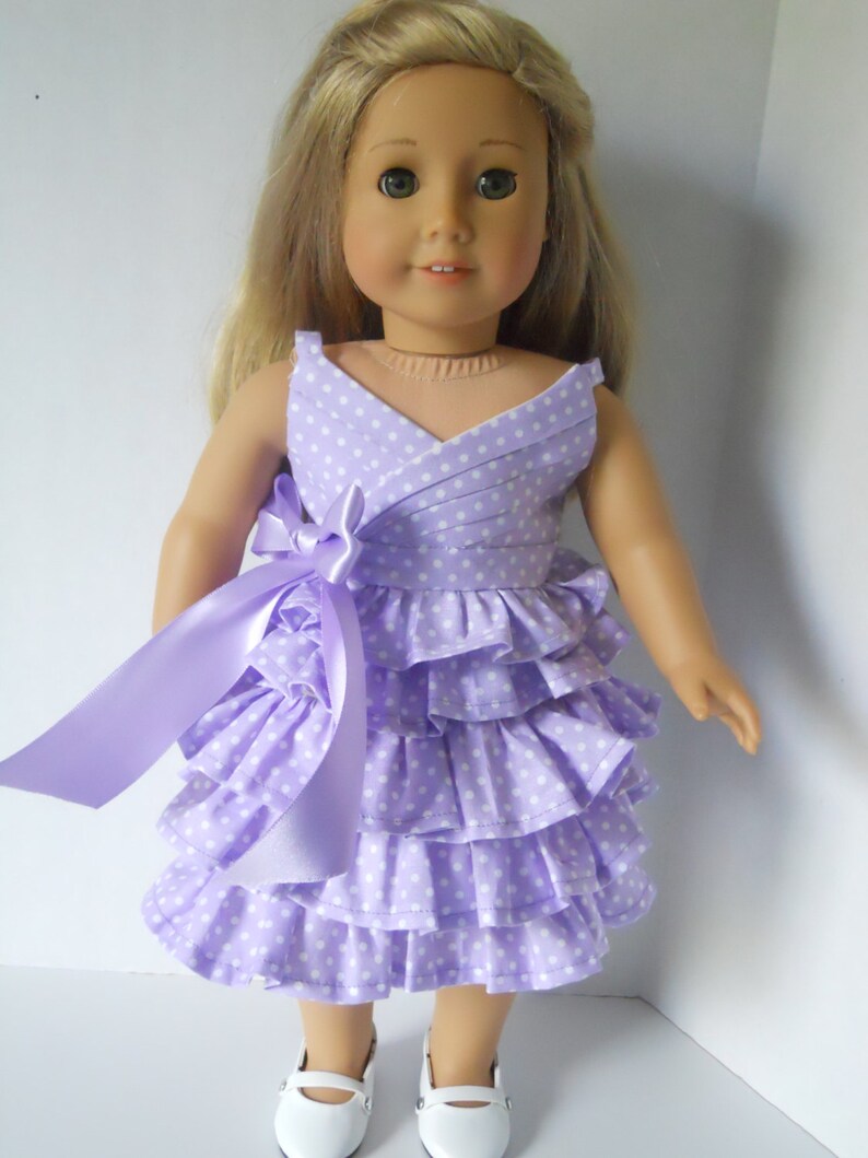 May include: A purple and white polka dot dress with a ruffled skirt and a matching purple ribbon. The dress is designed for a doll.