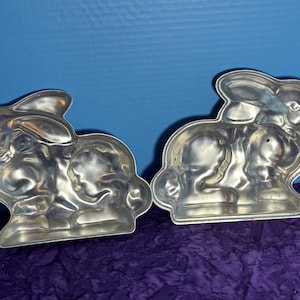 May include: Two silver metal bunny cake pans. The pans are in the shape of a bunny rabbit sitting up with its ears up.