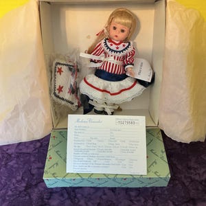 May include: A Madame Alexander doll in a box. The doll has blonde hair and wears a red, white, and blue dress with a star-studded collar and a white lace skirt. A small, star-adorned pillow is included. The box is green and white.