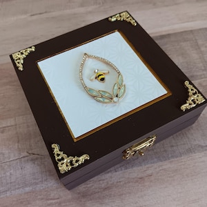 May include: A dark brown wooden box with gold-colored corner accents and a gold clasp. The box features a white interior with a gold-framed inset, showcasing a gold-toned teardrop-shaped pendant with a bee and leaf design.