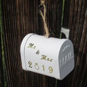May include: White metal mailbox ornament with the text "Mr & Mrs 2019" and "MAIL" on the side. The ornament is hanging from a wooden fence with twine.