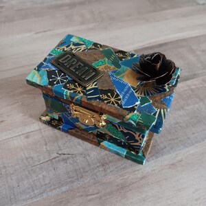 May include: A small wooden box with a blue, green, and gold patterned design. The box has a metal latch and a metal rose on top. The box has a metal tag on the front that says "DREAM".