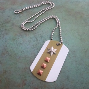 May include: A white and green dog tag necklace with a silver chain. The dog tag has a silver star and four copper pyramid studs.