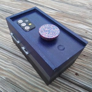 May include: A navy blue wooden box with two drawers and a matching lid. The lid features a black phone case with gear-shaped designs and a round, glitter-covered object. The box is on a wooden surface.