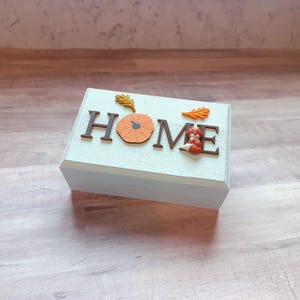 May include: A small wooden box with a light blue paint finish. The box has the word "HOME" spelled out in brown letters with a small orange pumpkin and a fox figurine in between the letters. The box is decorated with small orange and yellow leaves.