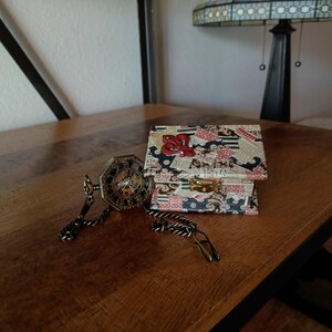 May include: A small, decorative box with a hinged lid. The box is covered in a patchwork of patterned paper with red, white, and black accents. A small, ornate metal keyhole is visible on the front of the box. A gold-toned pocket watch with a chain is lying on a wooden surface next to the box.