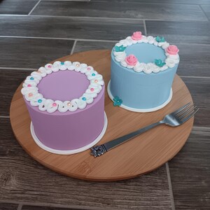 May include: Two small, round cake boxes with decorative frosting. The box on the left is purple with white frosting and colorful sprinkles. The box on the right is blue with white frosting and pink flowers. The boxes are on a wooden tray with a silver fork.
