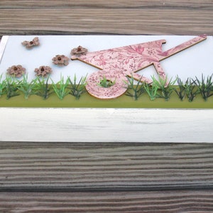 May include: A white wooden box with a light blue background. The box has a green grass border and a pink and white wheelbarrow with brown flowers on top.