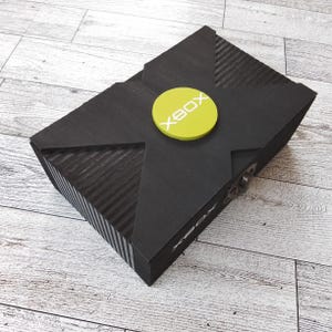 May include: A black wooden box with a green circle on the top that says "XBOX" in white letters. The box has a silver latch on the side.
