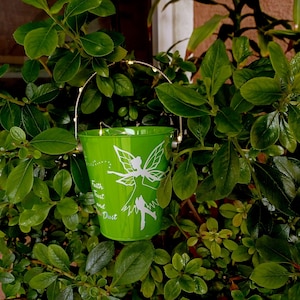 May include: A bright green metal bucket with a fairy design and the words "Faith Trust and Dust" in white. The bucket has a gold-colored handle and is surrounded by green leaves.