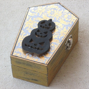 May include: A small wooden box shaped like a coffin with a gold and silver patterned design. The lid has a black silhouette of three stacked jack-o'-lanterns.