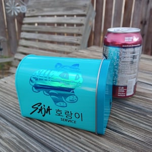 May include: A turquoise metal container with a cartoon tiger design and the text "Saja 호랑이 SERVICE". A red and silver soda can is in the background. The container is on a wooden surface.