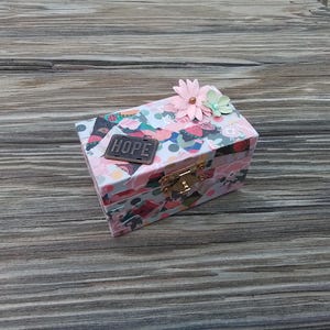 May include: A small, pink and green patterned box with a gold latch and a metal tag that says "HOPE". The box has a pink flower on top.