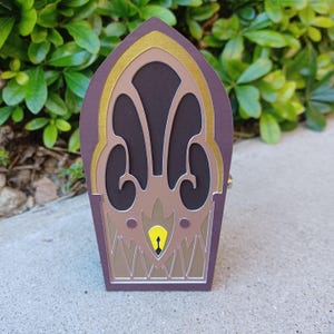 May include: A decorative, arched box with a dark purple exterior and a layered design. The box features gold, brown, and tan accents, with a keyhole detail at the bottom. The box is set against a backdrop of green foliage.